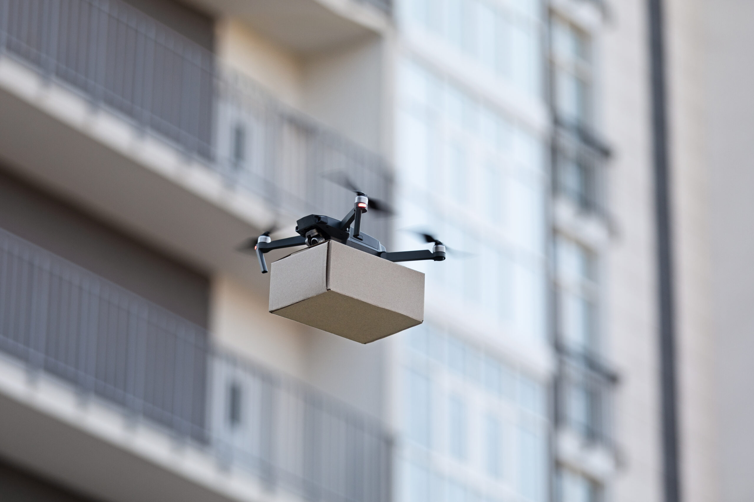 A drone in flight carrying a brown package flying in front of a city building.