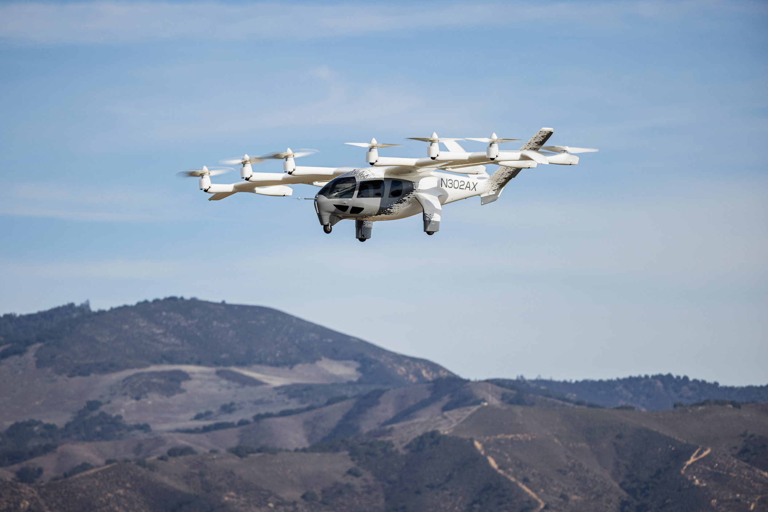 A Joby electric air taxi flying through a clear blue sky above a mountain range.