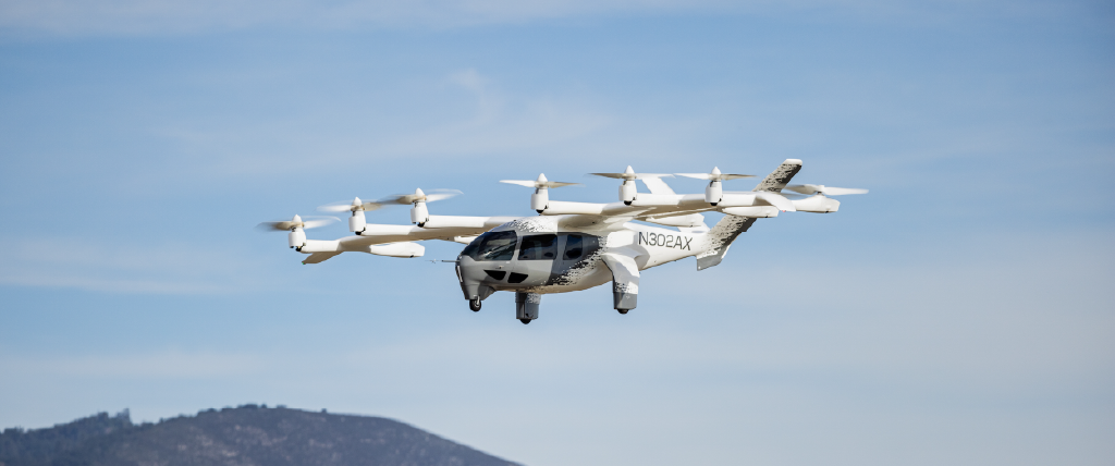 A Joby electric air taxi flying through a clear blue sky above a mountain range.