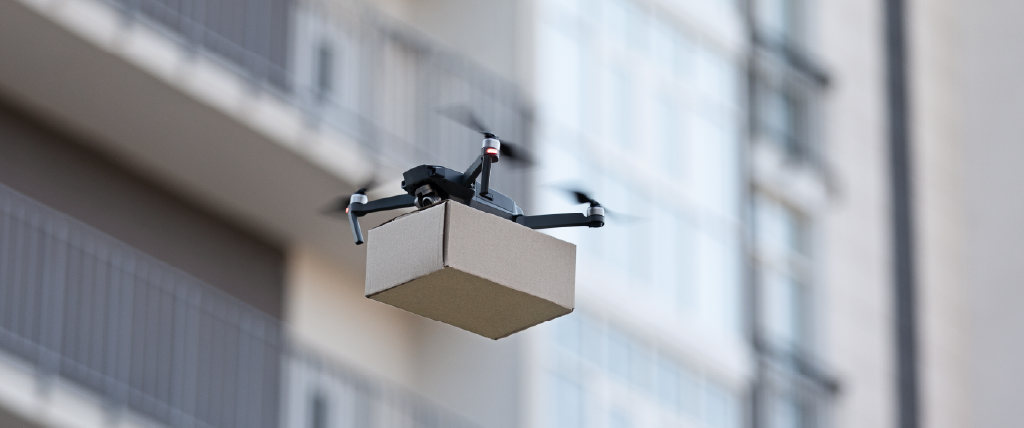 A drone in flight carrying a brown package flying in front of a city building.