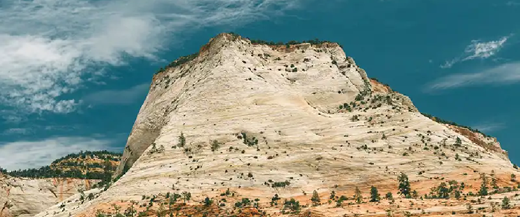 A large, light-colored sandstone mountain dominates the frame under a dramatic blue sky with wispy clouds. The mountain's face is a mix of off-white and light tan, marked by horizontal striations and dotted with small green trees and shrubs. The base of the mountain is a darker reddish-orange, suggesting a different rock layer or soil. To the left, another, darker mountain peak is partially visible. The overall impression is one of grandeur and rugged natural beauty.