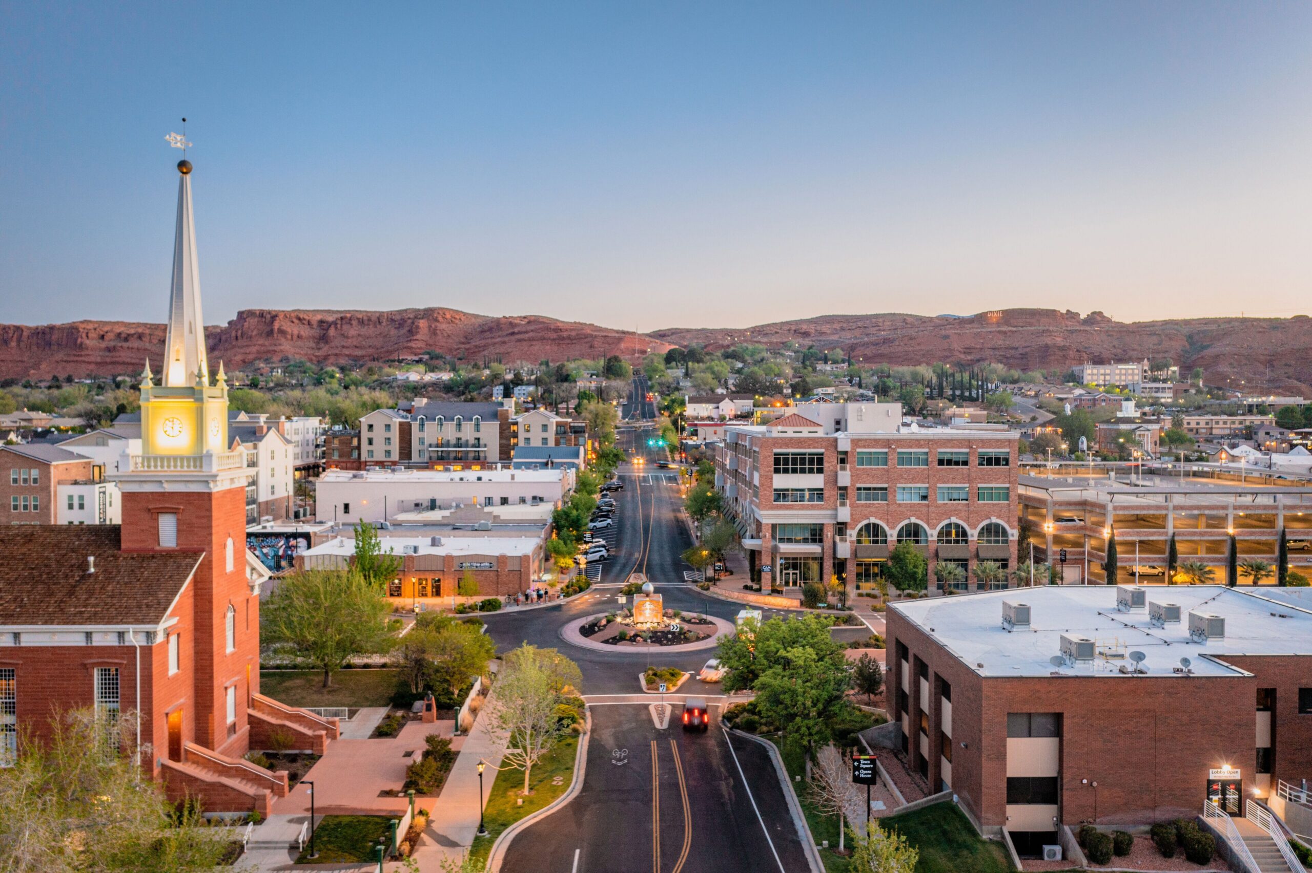 The city of St. George, Utah at dusk, with the red rock mountains in the background. There are historic buildings and a roundabout in the center.