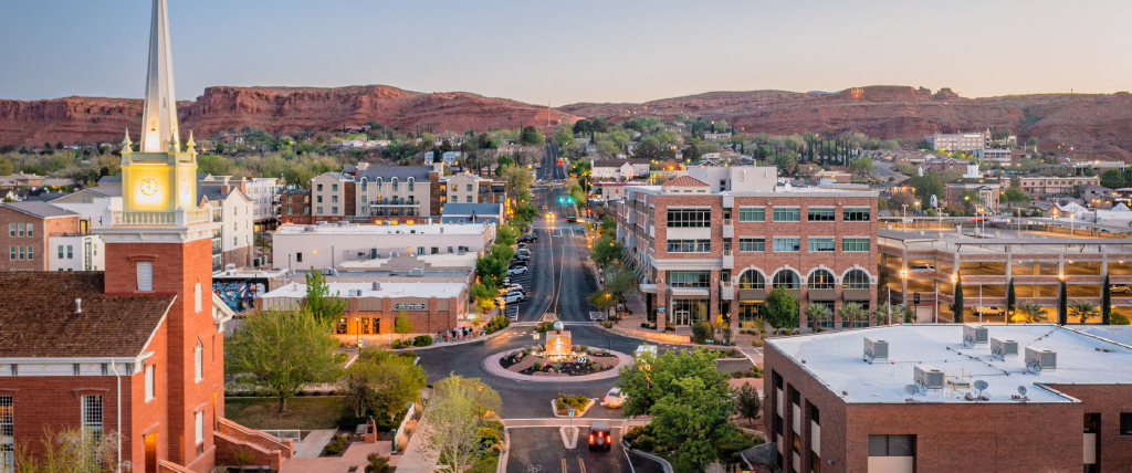 The city of St. George, Utah at dusk, with the red rock mountains in the background. There are historic buildings and a roundabout in the center.